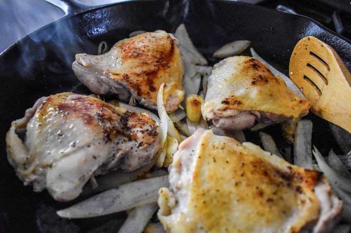 chicken thighs cooking in a cast iron skillet with onion and garlic.