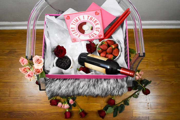 bottle of red wine, strawberries, flowers, chocolate, candles, and Valentine's Day cards in a box sitting on a ghost chair