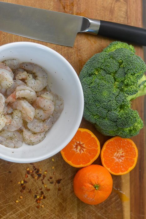 seasoned raw shrimp in white bowl, crown of broccoli, oranges, and red pepper flakes on wooden cutting board.