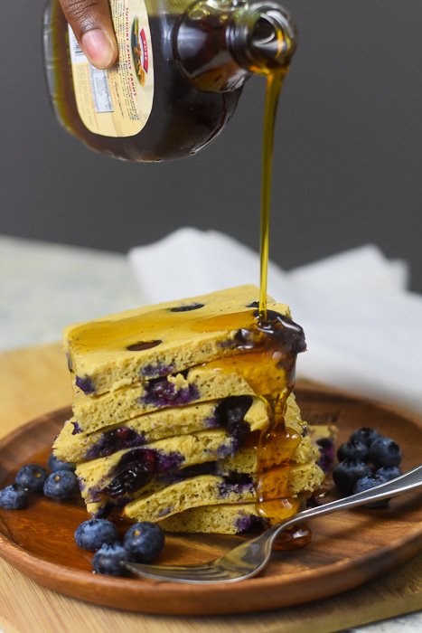 pouring maple syrup on stack of blueberry protein pancakes.