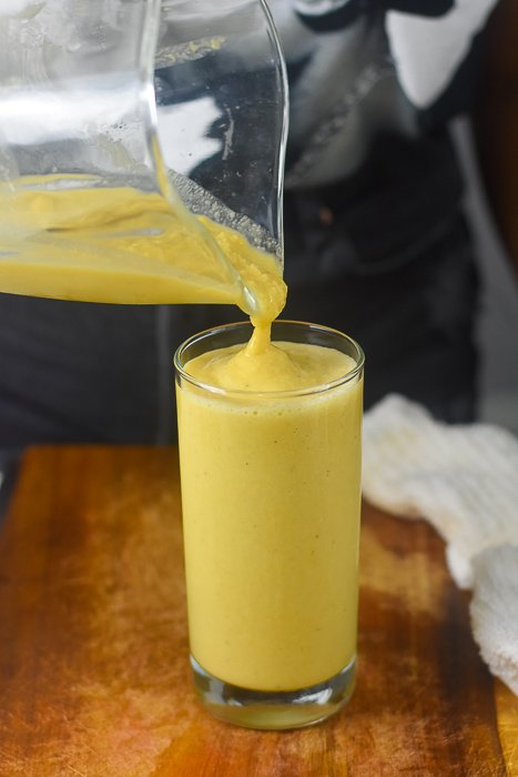 pouring golden pineapple smoothie into tall, clear glass.