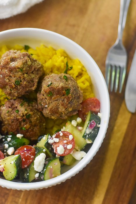 white bowl filled with cucumber Greek salad, yellow rice, and lamb meatballs.