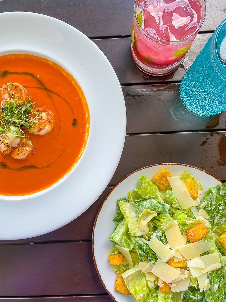 bowl of tomato soup, cocktail, and Caesar salad on table