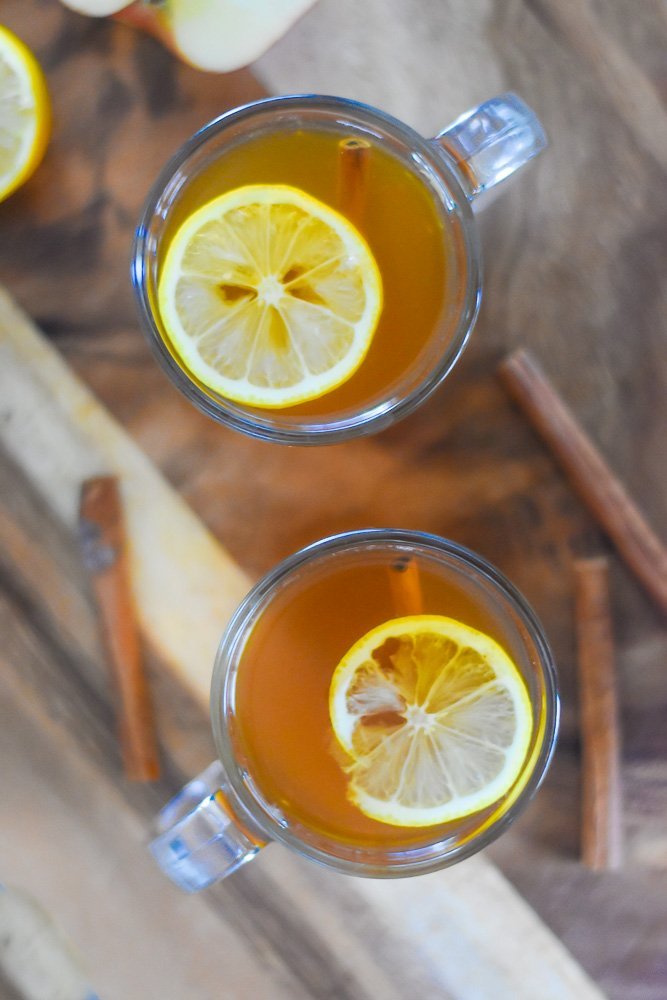 lemon slices floating in two clear mugs of cider hot toddies on wooden cutting board.