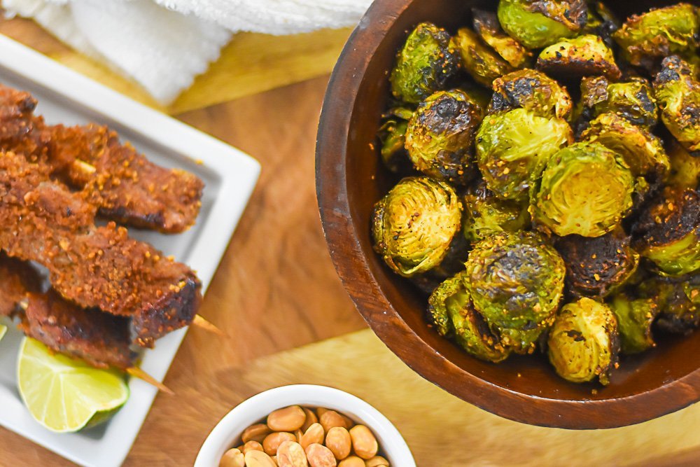 roasted brussels sprouts in wooden bowl next to plate of beef suya skewers.