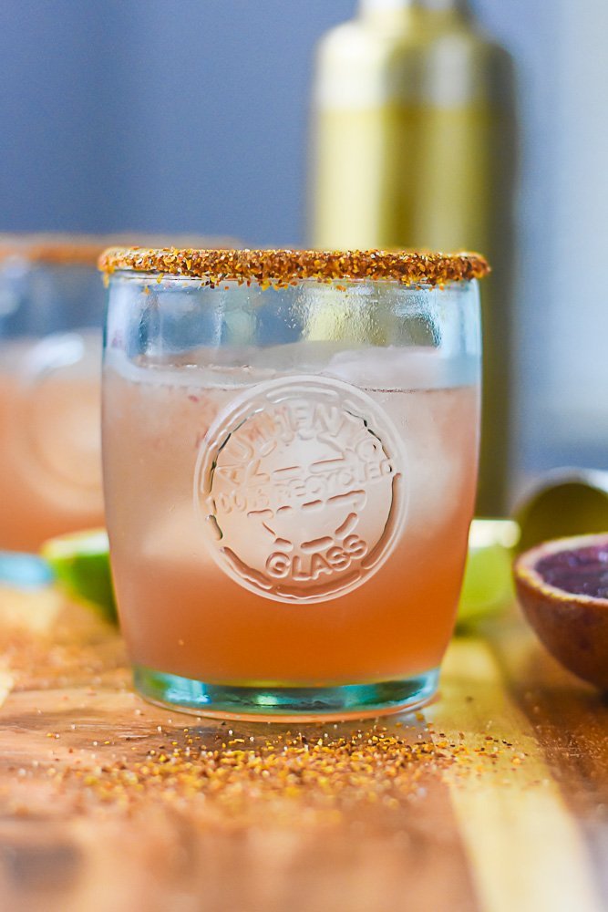 blood orange mezcal margarita in recycled glass cup surrounded by sprinkled tajín seasoning, fresh blood oranges and limes.