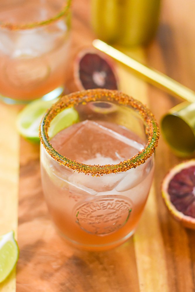two blood orange mezcal margaritas surrounded by fresh blood oranges and lime wedges.