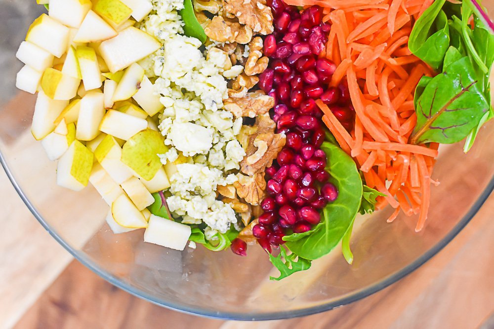 cut pears, gorgonzola cheese, walnut pieces, pomegranate arils, matchstick carrots, and super greens blend in large glass bowl.