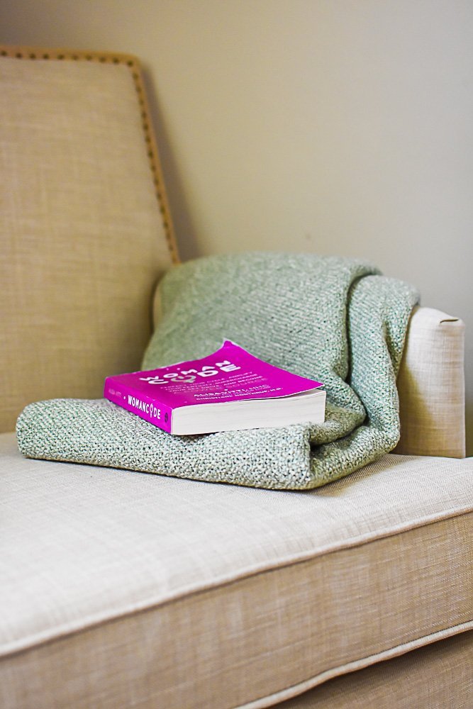 green throw blanket and pink paperback book on seat of reading chair.