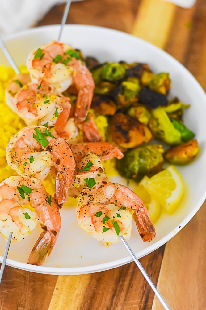 two skewers of lemon pepper shrimp over brussels sprouts and yellow rice on a white plate.