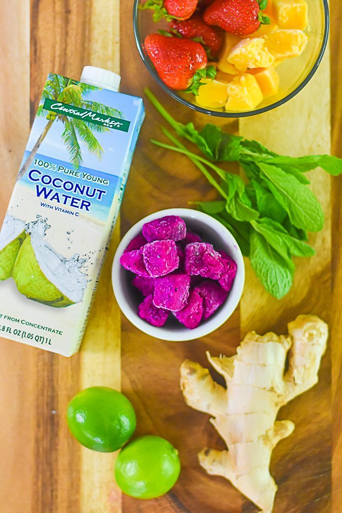 carton of coconut water, glass bowl of mango chunks and strawberries, fresh mint sprigs, bowl of frozen dragon fruit chunks, fresh ginger, and two limes on wooden cutting board.