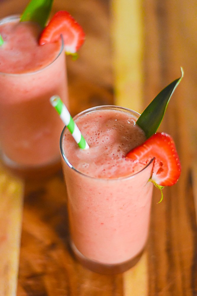 two glasses of frosé garnished with fresh strawberry slices, pineapple fronds, and green striped paper straws.