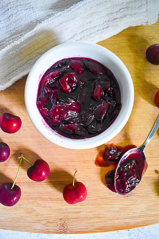 small bowl of cherry jam surrounded by fresh cherries on wooden cutting board.