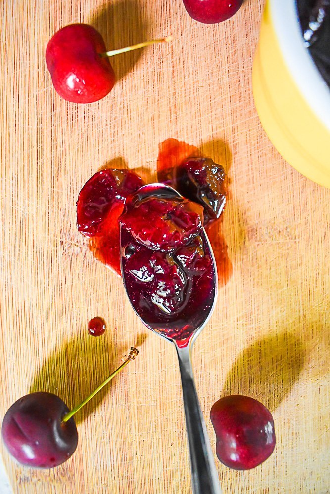 spoon of cherry jam surrounded by fresh cherries on wooden board.