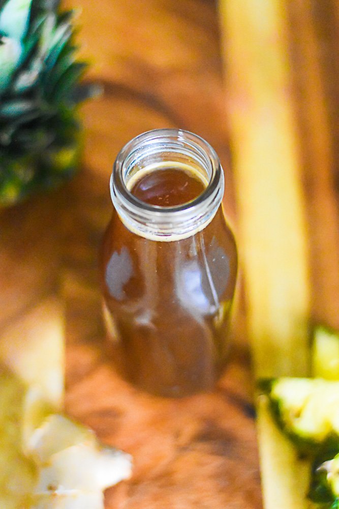 spiced pineapple syrup made with brown sugar in a glass container on wooden cutting board surrounded by fresh pineapple and ginger pieces.