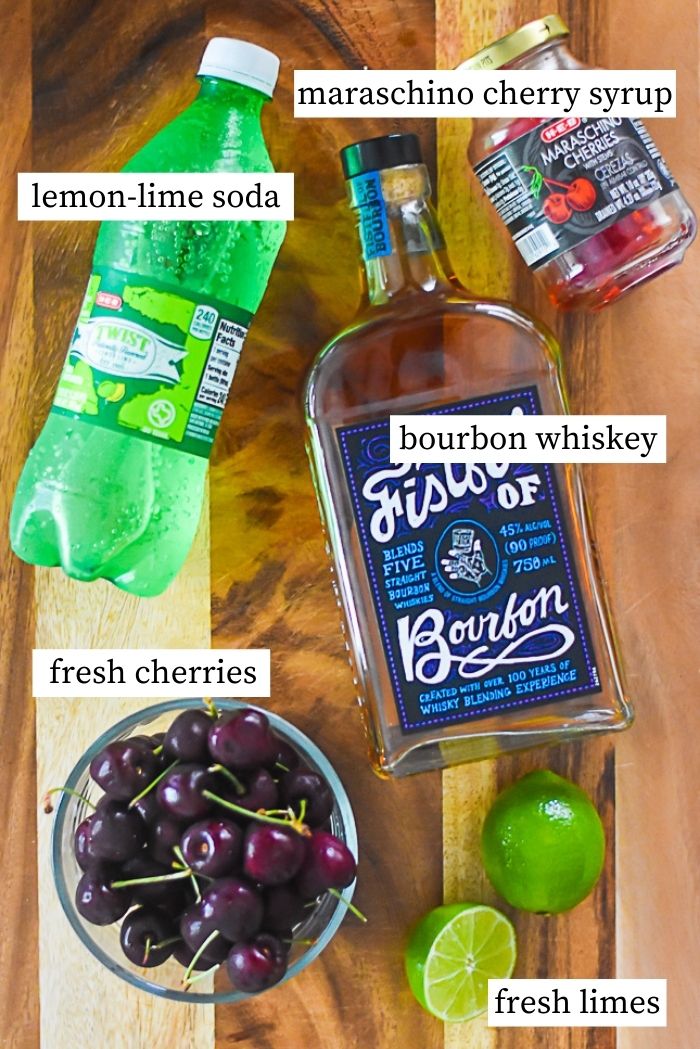 flat lay of ingredients for bourbon cherry limeade: bowl of fresh cherries, fresh limes, bottle of bourbon, bottle of lemon lime soda, jar of maraschino cherry syrup.