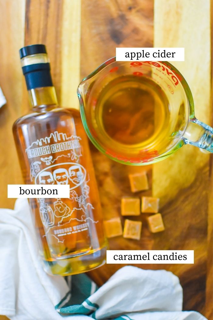 flat lay of Brough Brothers Bourbon bottle, wrapped caramel candies, and measuring cup of apple cider on wooden counter.