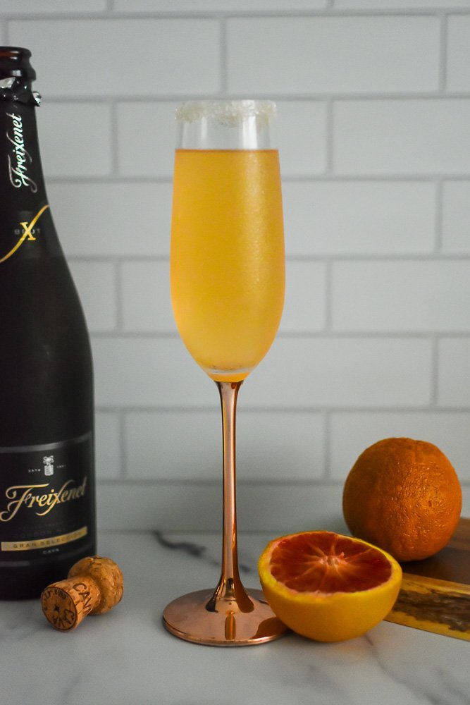 citrus champagne cocktail next to bottle of Freixenet sparkling wine, cork, and blood oranges.