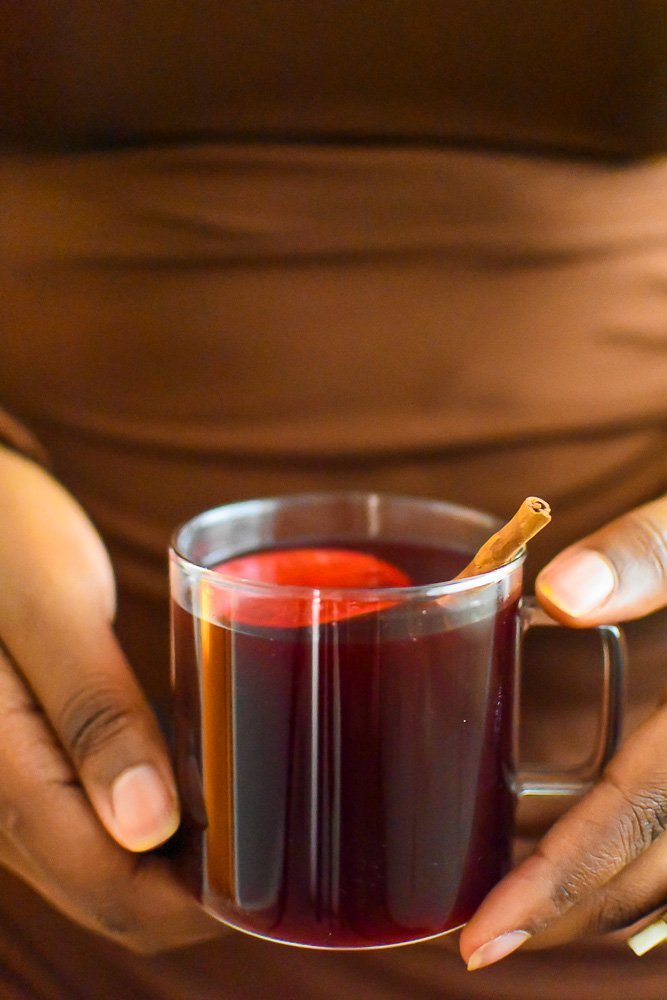 holding clear mug of hot zobo drink with cinnamon stick garnish.
