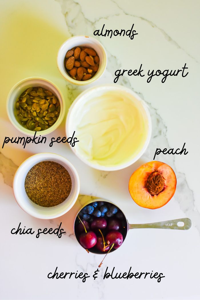 almonds, pumpkin seeds, greek yogurt, chia seeds, peach, blueberries, and cherries on granite counter.