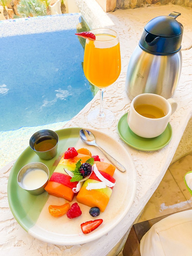 colorful plate of fresh fruit, yogurt and syrup next to mug of tea on green saucer and orange juice in wine glass on ledge of a private pool.