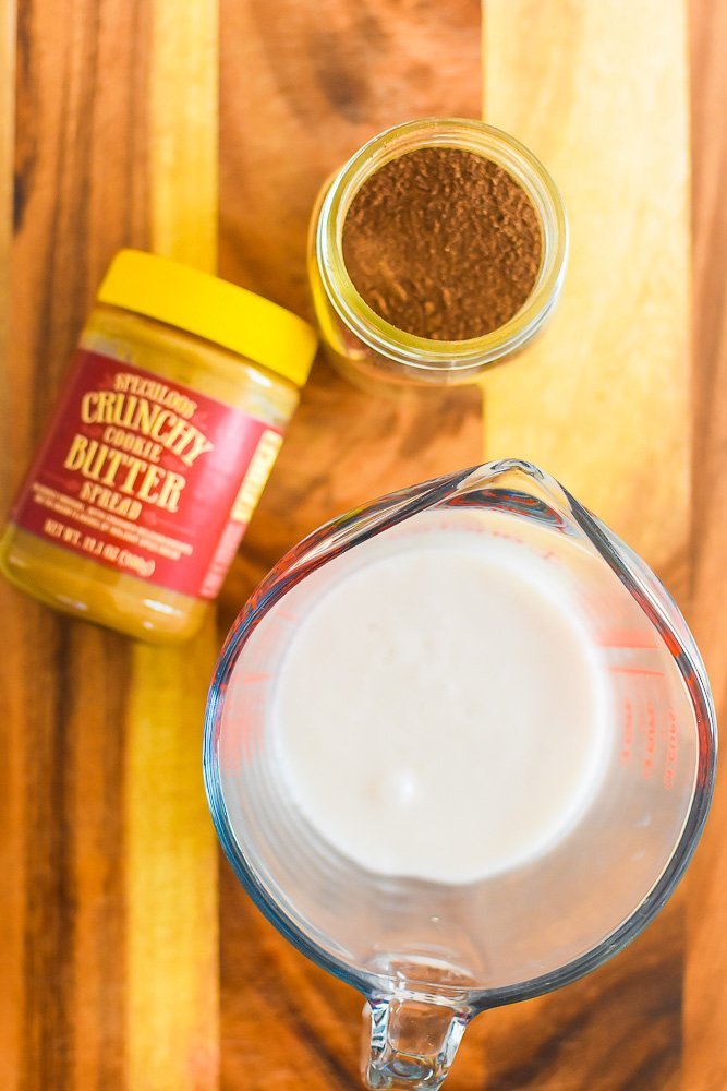 jar of cookie butter, jar of instant chai, and cup of steamed milk on wooden cutting board.