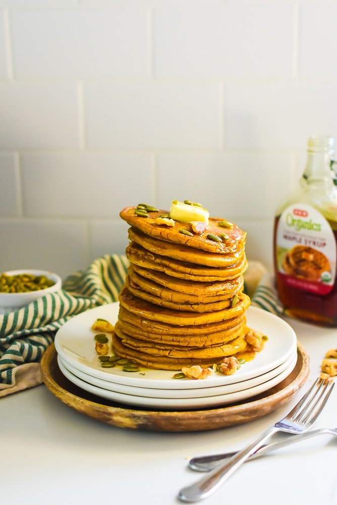 tall stack of PCOS friendly pumpkin pancakes served with maple syrup, pumpkin seeds, and walnuts.