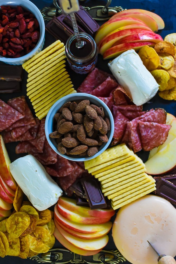 cheese board full of flavored almonds, peppered salami, logs of goat cheese, sliced honeycrisp apples, buttery crackers, jar of honey, plantain chips, and pieces of dark chocolate.