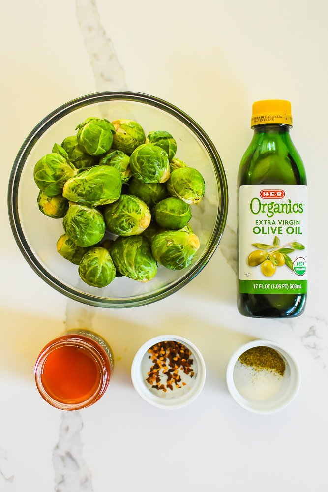 bottle of olive oil and bowls of raw brussels sprouts, honey, red pepper flakes, salt and pepper on granite counter top.