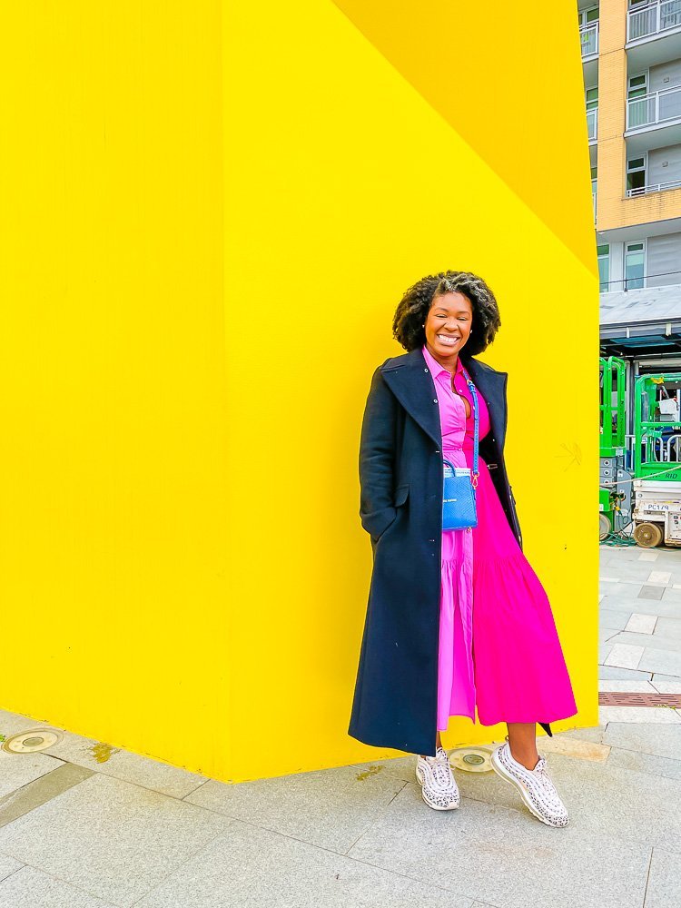 Jazzmine in long bright pink dress, black coat, and leopard sneakers standing in front of bright yellow wall outdoors.