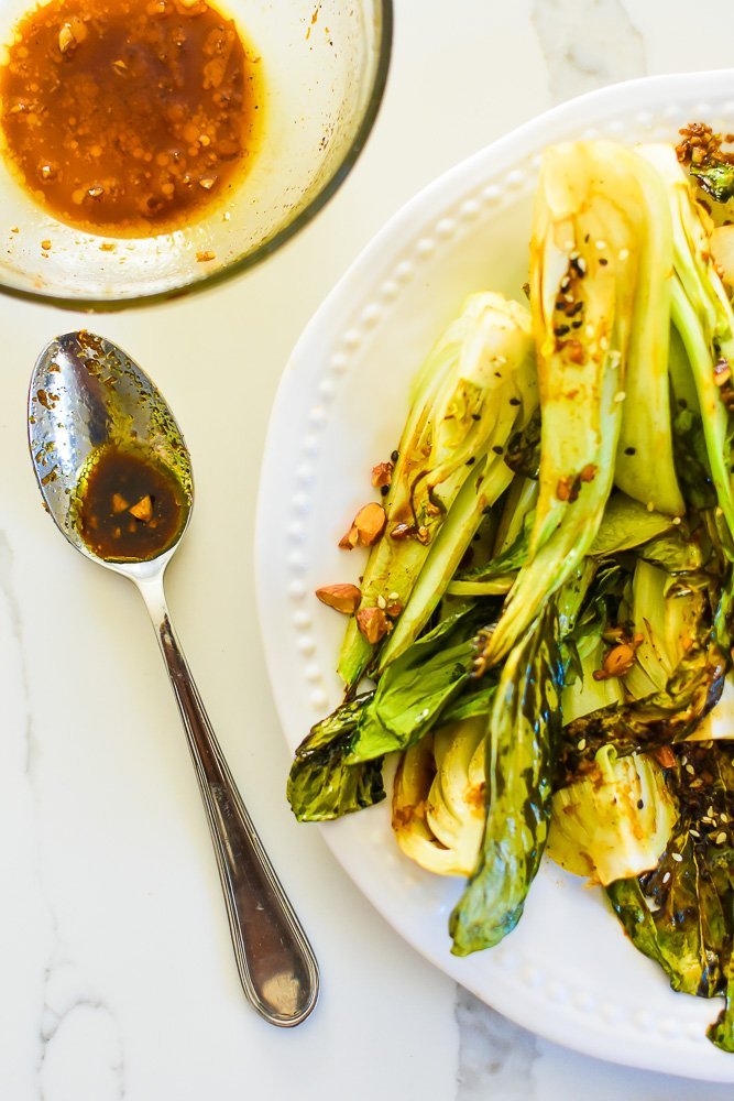 bowl and spoon of garlic soy sauce next to plate of roasted and garnished bok choy.