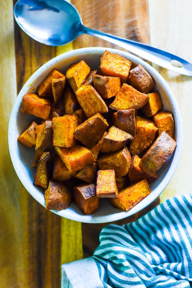 bowl of roasted sweet potatoes on countertop next to sriped kitchen towel and metal serving spoon.