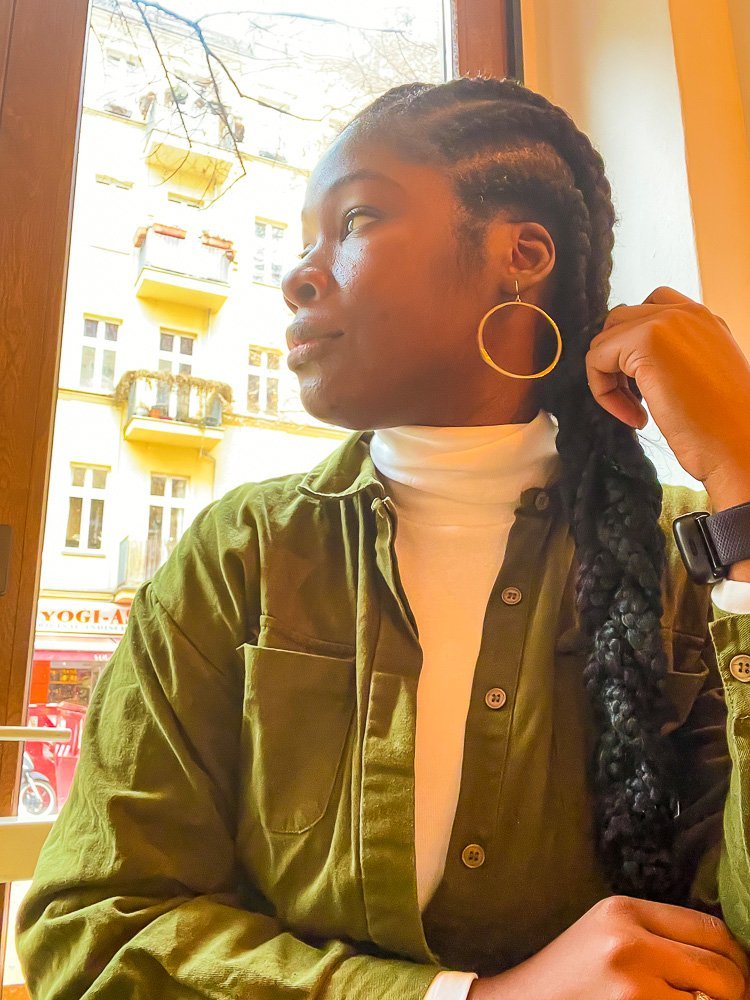 Jazzmine sitting next to a window in a Berlin coffee shop wearing a green flight suit and white turtleneck, gold hoop earrings, and her cornrows braided to the side.