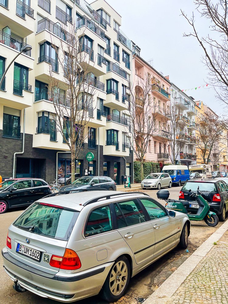 street view of a Berlin neighborhood including multi-story apartment buildings painted different colors and cars parallel parked on the street.