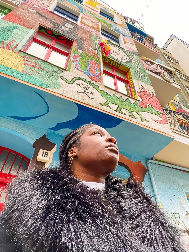Jazzmine standing in front of a brightly painted building wearing a dramatic furry collared coat.