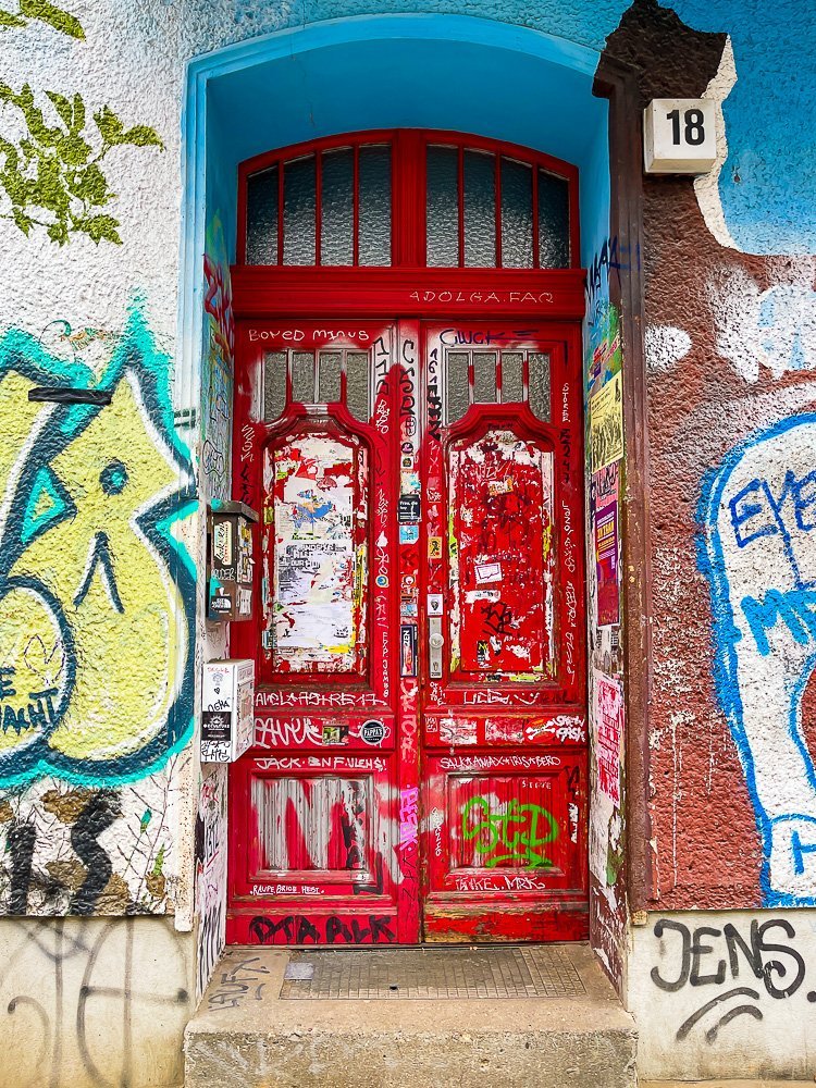 entryway in Berlin neighborhood with graffiti and stickers all over, included on the red double doors.