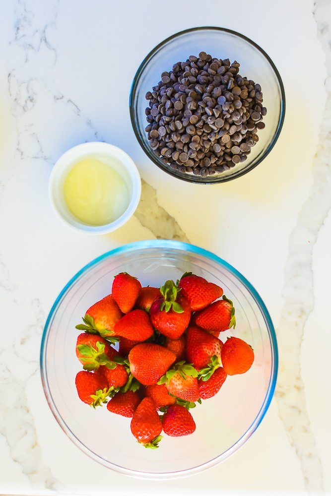 bowls of fresh strawberries, chocolate chips, and coconut oil on granite counter.