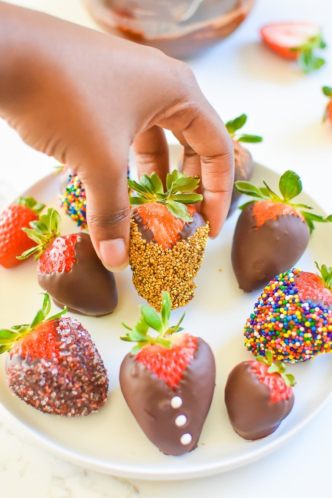 brown hand placing chocolate strawberry with gold sprinkles on round platter.