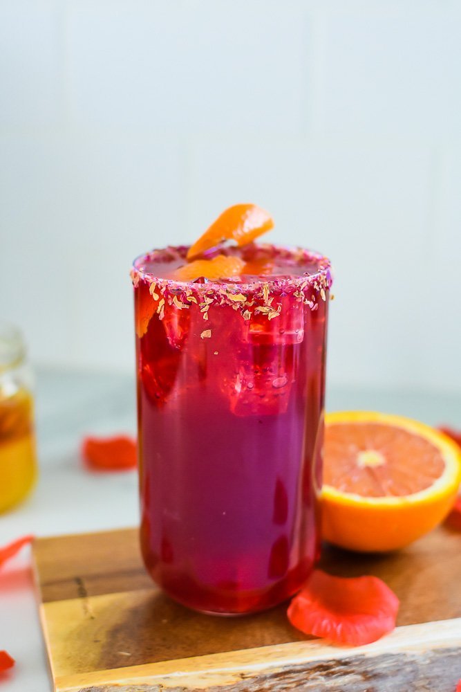 hibiscus flower mocktail on wooden cutting board with halved orange and rose petals scattered around.