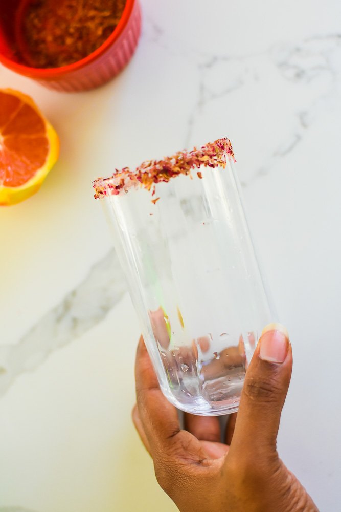 brown hand holding beveled highball glass rimmed in sugar and dried rose petals.
