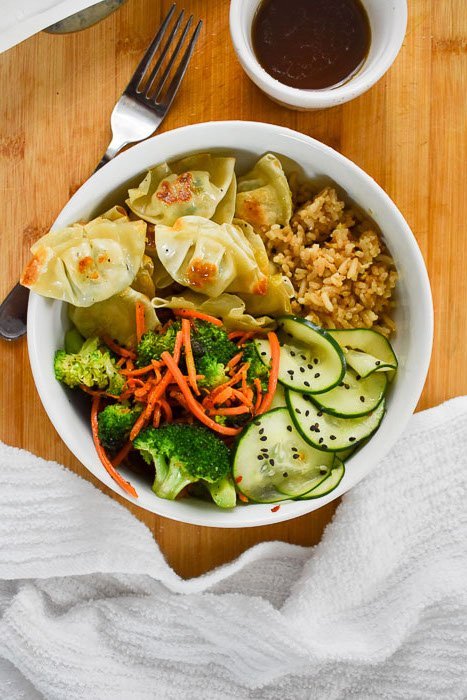 power bowl of brown rice, mini wontons, pickled cucumbers, and sauteed vegetables next to small bowl of brown finishing sauce on wooden cutting board.