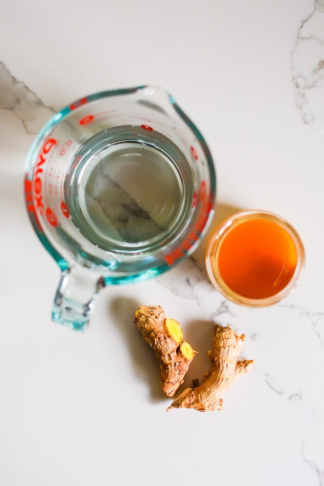 cup of water, jar of honey, and two pieces of fresh ginger on granite counter top.