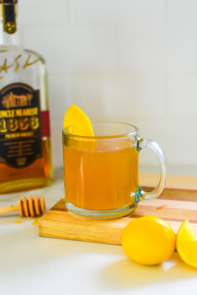 mint hot toddy in glass mug on wooden cutting board surrounded by fresh lemons, honey, and bottle of Uncle Nearest 1856 whiskey.