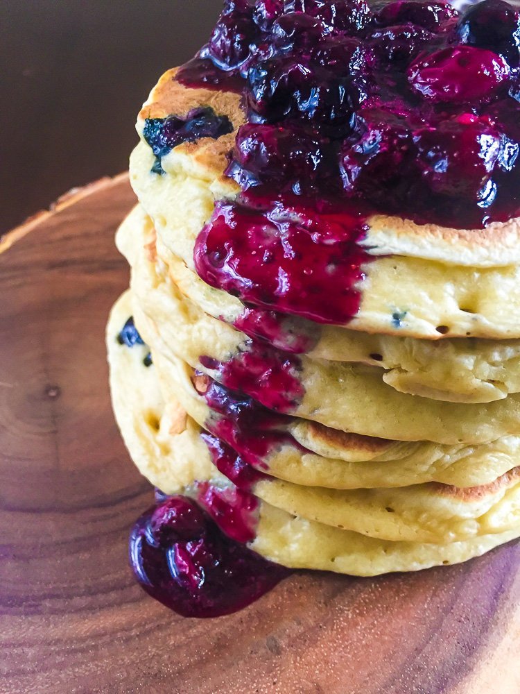 six pancakes stacked on top of each other and topped with blueberry compote sauce cascading down the side.