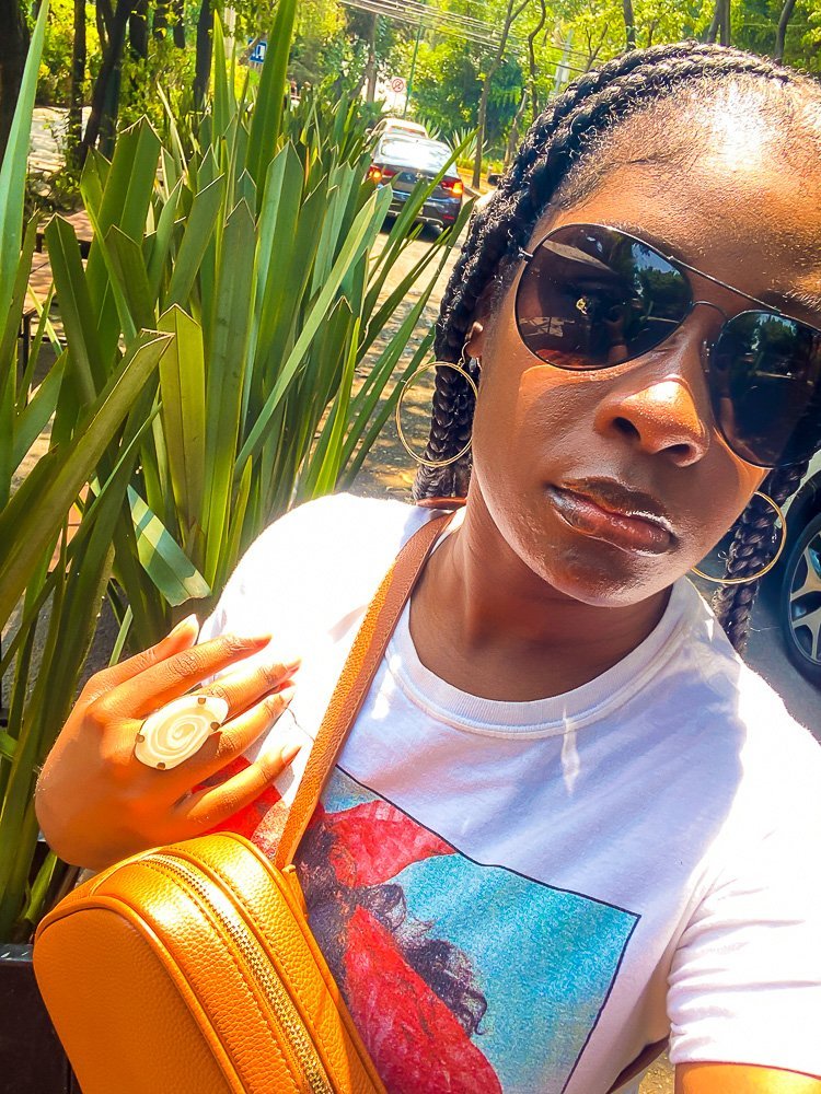 Close up selfie of Jazzmine wearing a graphic tee shirt, camel colored bum bag, hoop earrings, and black sunglasses.