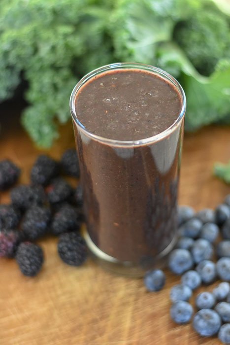açai, blueberry, blackberry, kale smoothie in a glass surrounded by fresh berries and bunch of kale.