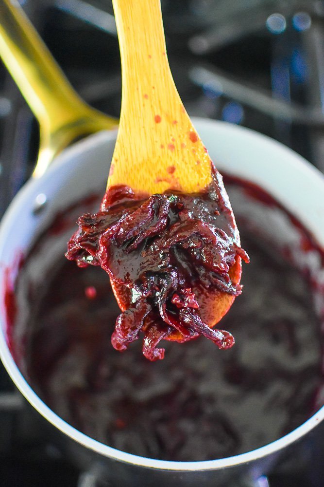 straining dried hibiscus flowers out of homemade BBQ sauce with wooden slotted spoon.