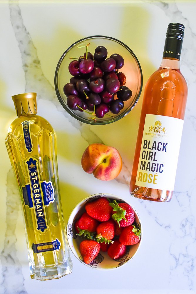 bowls of fresh cherries and strawberries, whole peach, and bottles of St. Germain elderflower liqueur and 
 Black Girl Magic rosé wine on granite countertop.