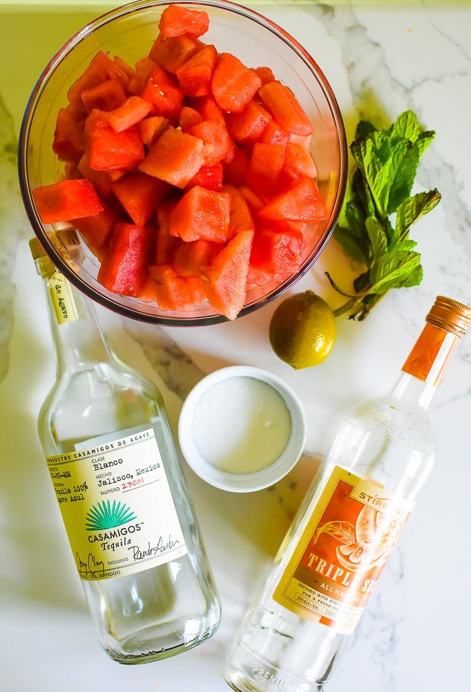 fresh watermelon chunks, fresh mint, bottles of tequila and orange liqueur, sugar, and a lime on granite countertop.