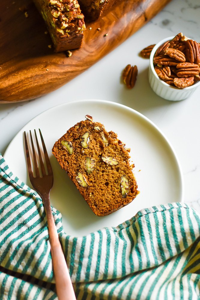slice of banana nut bread with pecans on white plate with copper fork and striped towel next to it.